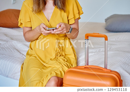 Woman in dress sitting on bed with orange suitcase using smartphone indoors Woman in dress sitting on bed with orange suitcase using smartphone indoors 129453118