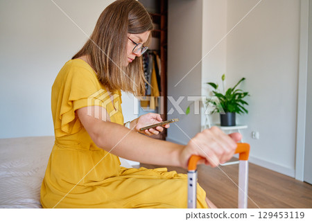 Woman sitting on bed with suitcase and using smartphone Woman sitting on bed with suitcase and using smartphone 129453119