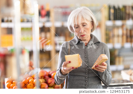 Choose cheese in supermarket - elderly woman chooses what cheese to buy in the dairy department section of store 129453175