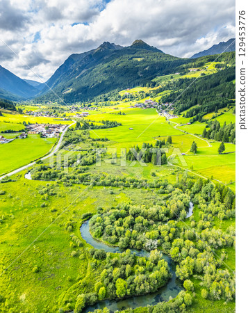 A winding river flows gracefully through a vibrant green valley surrounded by majestic mountains in the Alps. The landscape is dotted with patches of trees and meadows, showcasing nature's beauty. A winding river flows gracefully through a vibrant green valley surrounded by majestic mountains in the Alps. The landscape is dotted with patches of trees and meadows, showcasing nature's beauty. 129453776