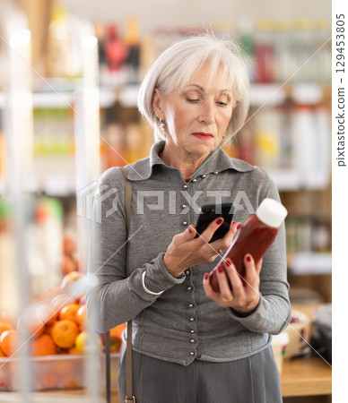 Elderly female shopper scanning QR code on bottle ketchup label in grocery department of supermarket 129453805