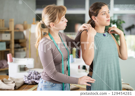 Young tailoress and female client looking ready dress in sewing workshop Young tailoress and female client looking ready dress in sewing workshop 129454281