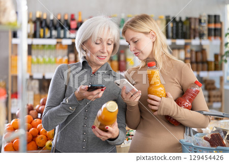 Elderly mother and her adult daughter scan a QR code on a tomato and peach juice label together to check expiration date 129454426