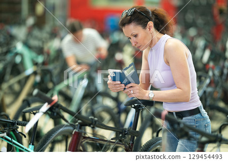 Positive woman buyer using smartphone for examining quality seat and choosing bike in bicycle sports store Positive woman buyer using smartphone for examining quality seat and choosing bike in bicycle sports store 129454493