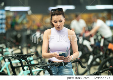 Happy woman buying new bike in bicycle sports store, using smartphone to scan barcode of products Happy woman buying new bike in bicycle sports store, using smartphone to scan barcode of products 129454573