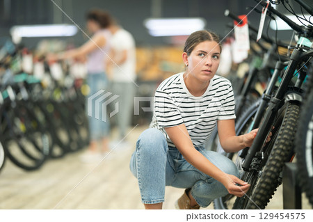 Glad young girl selects bicycle in sporting goods store, checking wheel quality Glad young girl selects bicycle in sporting goods store, checking wheel quality 129454575