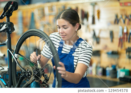 Professional young girl master wearing uniform fixing bike, lubricating bicycle chain in sport store Professional young girl master wearing uniform fixing bike, lubricating bicycle chain in sport store 129454578