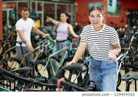 Smiling girl assessing saddle while choosing bicycle in store 129454592
