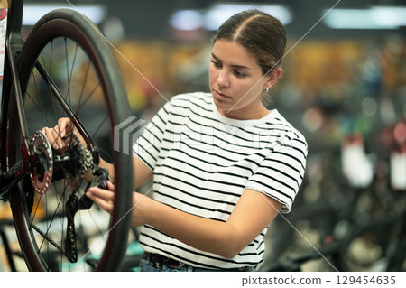 Attentive young female buyer examining bicycle wheel in store Attentive young female buyer examining bicycle wheel in store 129454635