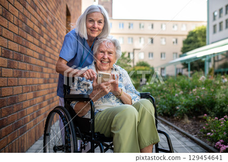 Senior patient in wheelchair showing picture in smartphone to caregiver. Senior patient in wheelchair showing picture in smartphone to caregiver. 129454641