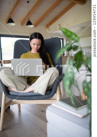 Woman working on laptop in cozy living room, sitting cross-legged on armchair. Woman working on laptop in cozy living room, sitting cross-legged on armchair. 129454731