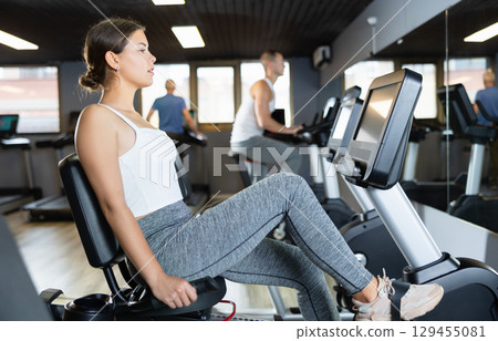 Concentrated young girl working out on stationary bicycle in gym. Healthy active lifestyle concept Concentrated young girl working out on stationary bicycle in gym. Healthy active lifestyle concept 129455081