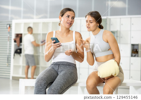 In locker room of sports club, girl communicates with female friend holding phone in her hands. In locker room of sports club, girl communicates with female friend holding phone in her hands. 129455146