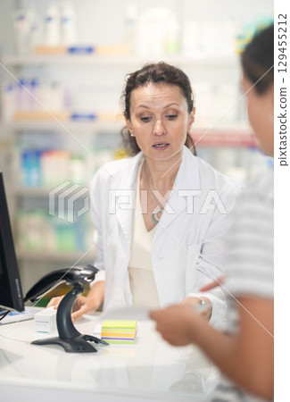 Female pharmacist at cash register scans a QR code and sells medicine to pharmacy visitor. 129455212