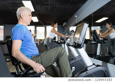 Elderly man, along with other people, is engaged on an bike in gym Elderly man, along with other people, is engaged on an bike in gym 129455383