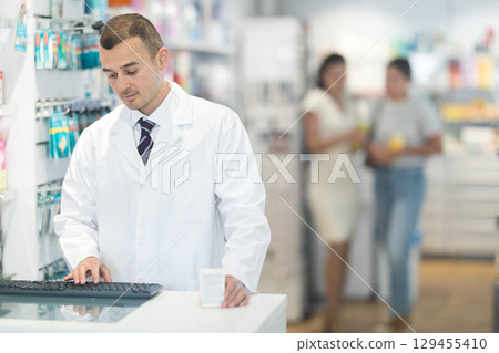 Young male pharmacist standing at the desk using keyboard in chemist's shop Young male pharmacist standing at the desk using keyboard in chemist's shop 129455410