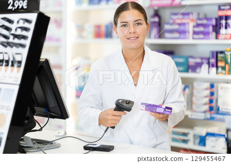 Girl pharmacist in cash area works with computer and goods, scans drugs, checks availability 129455467