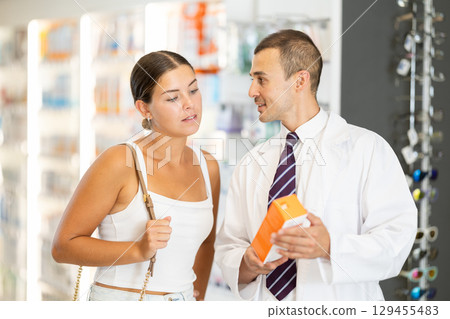 Male consultant pharmacist holds cream in hands and offers girl client to purchase Male consultant pharmacist holds cream in hands and offers girl client to purchase 129455483