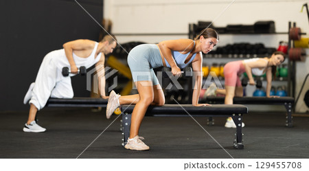 Athletic young woman pumping her biceps with a dumbbell while leaning on bench Athletic young woman pumping her biceps with a dumbbell while leaning on bench 129455708