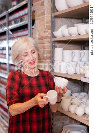 Elderly woman chooses mug or cup in pottery and ceramics shop 129456014