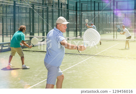 Mature man playing padel game in court on sunny day Mature man playing padel game in court on sunny day 129456016
