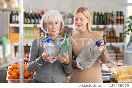 Girl with grandmother buyers examine goods, choose large capacity bottle of water in store 129456079