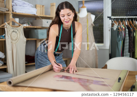 Positive young female dressmaker drawing on paper in sewing workshop Positive young female dressmaker drawing on paper in sewing workshop 129456117