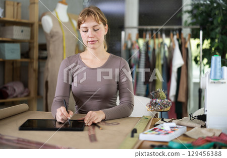 Young female dressmaker drawing in tablet while sitting at table 129456388