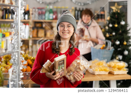 Smiling girl making purchases in food department of supermarket, choosing tasty sweets for celebration of Christmas 129456411