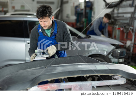 Male mechanic in uniform polishes bumper of car with machine in car service station 129456439