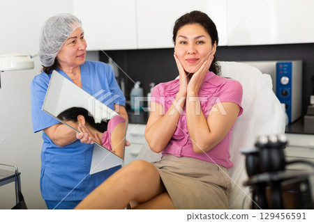 Portrait of a satisfied young female patient after the procedure, looking in the mirror Portrait of a satisfied young female patient after the procedure, looking in the mirror 129456591