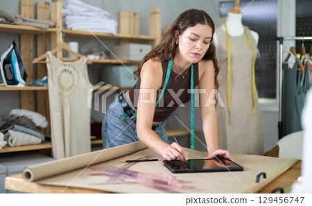 Young female dressmaker drawing in tablet in sewing workshop Young female dressmaker drawing in tablet in sewing workshop 129456747