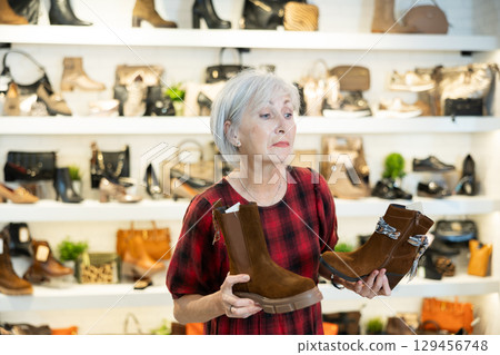 Elderly woman buyer looks at footwear from new collection and picks up new pair of boots for winter. Elderly woman buyer looks at footwear from new collection and picks up new pair of boots for winter. 129456748