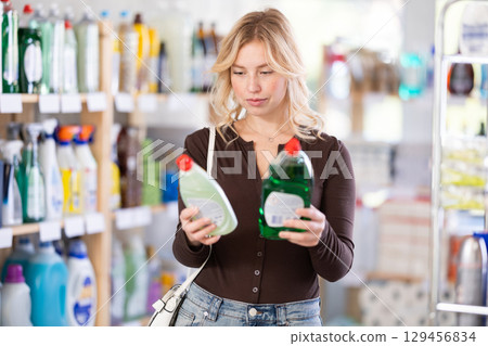 Attentive young Woman choosing between two dishwashing liquids standing between shelves in supermarket 129456834