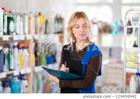 Beautiful diligent young girl saleswoman standing between shelves and doing inventory in hypermarket Beautiful diligent young girl saleswoman standing between shelves and doing inventory in hypermarket 129456942
