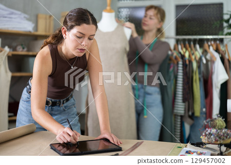 Young female dressmaker drawing in tablet other work with mannequin 129457002