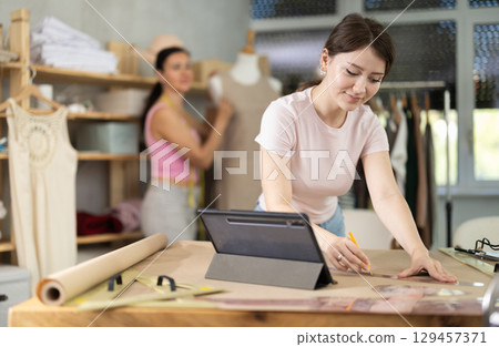 Girl tailor draws dress pattern of product on paper, tablet on table. Companion in background 129457371