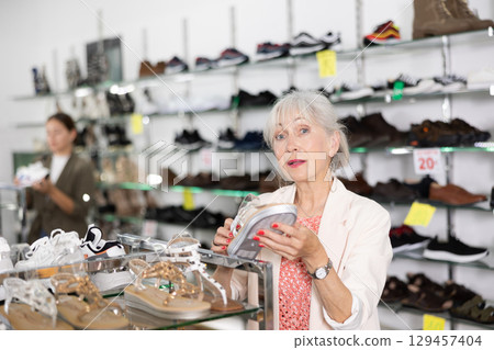Mature woman who came to shoe store for shopping, chooses summer shoes 129457404