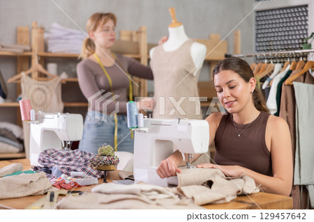 Woman tailor working on a sewing machine with an assistant in the background Woman tailor working on a sewing machine with an assistant in the background 129457642