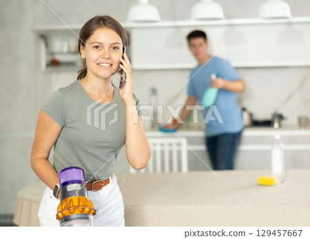 Young woman vacuuming the floor while talking on phone and her boyfriend dusting in the kitchen 129457667