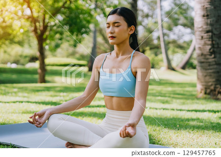 Asian woman meditating in a peaceful park setting, sitting cross-legged on green grass Asian woman meditating in a peaceful park setting, sitting cross-legged on green grass 129457765