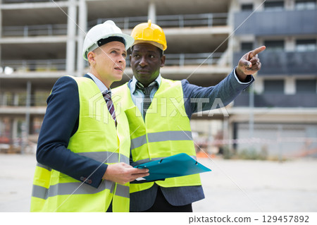 African-american and European men standing on construction site and talking. One of them holding folder with documentation, another one showing something with finger gesture. African-american and European men standing on construction site and talking. One of them holding folder with documentation, another one showing something with finger gesture. 129457892