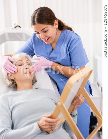 Young woman doctor cosmetologist holding mirror and showing result of mesotherapy and fillers with hyaluronic acid to mature woman in clinic 129458075