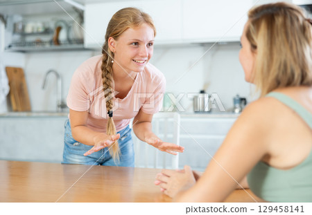 Cheerful teenage daughter talking friendly to mother in kitchen Cheerful teenage daughter talking friendly to mother in kitchen 129458141