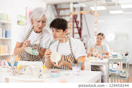 Elderly female teacher assisting guy painting ceramic mug in pottery class Elderly female teacher assisting guy painting ceramic mug in pottery class 129458244