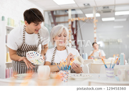 Young ceramicist teaching elderly woman to paint mug in pottery studio Young ceramicist teaching elderly woman to paint mug in pottery studio 129458360