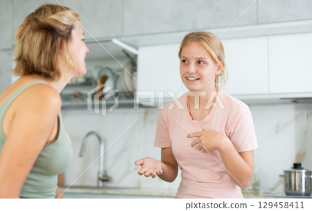 Cheerful teenage daughter talking friendly to mother in kitchen Cheerful teenage daughter talking friendly to mother in kitchen 129458411