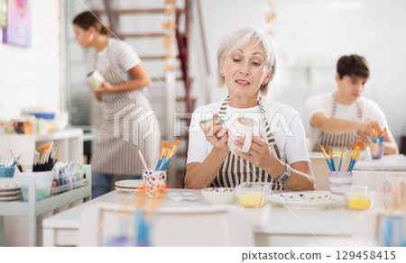 Talented senior female artisan applying design to ceramic mug in pottery studio 129458415