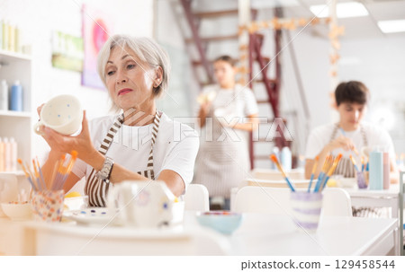 Interested elderly woman painting on ceramic cup during pottery class Interested elderly woman painting on ceramic cup during pottery class 129458544