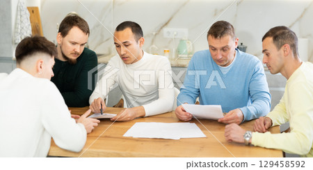 Friends reading financial documents and trying to solve problems together while sitting at table in their kitchen Friends reading financial documents and trying to solve problems together while sitting at table in their kitchen 129458592
